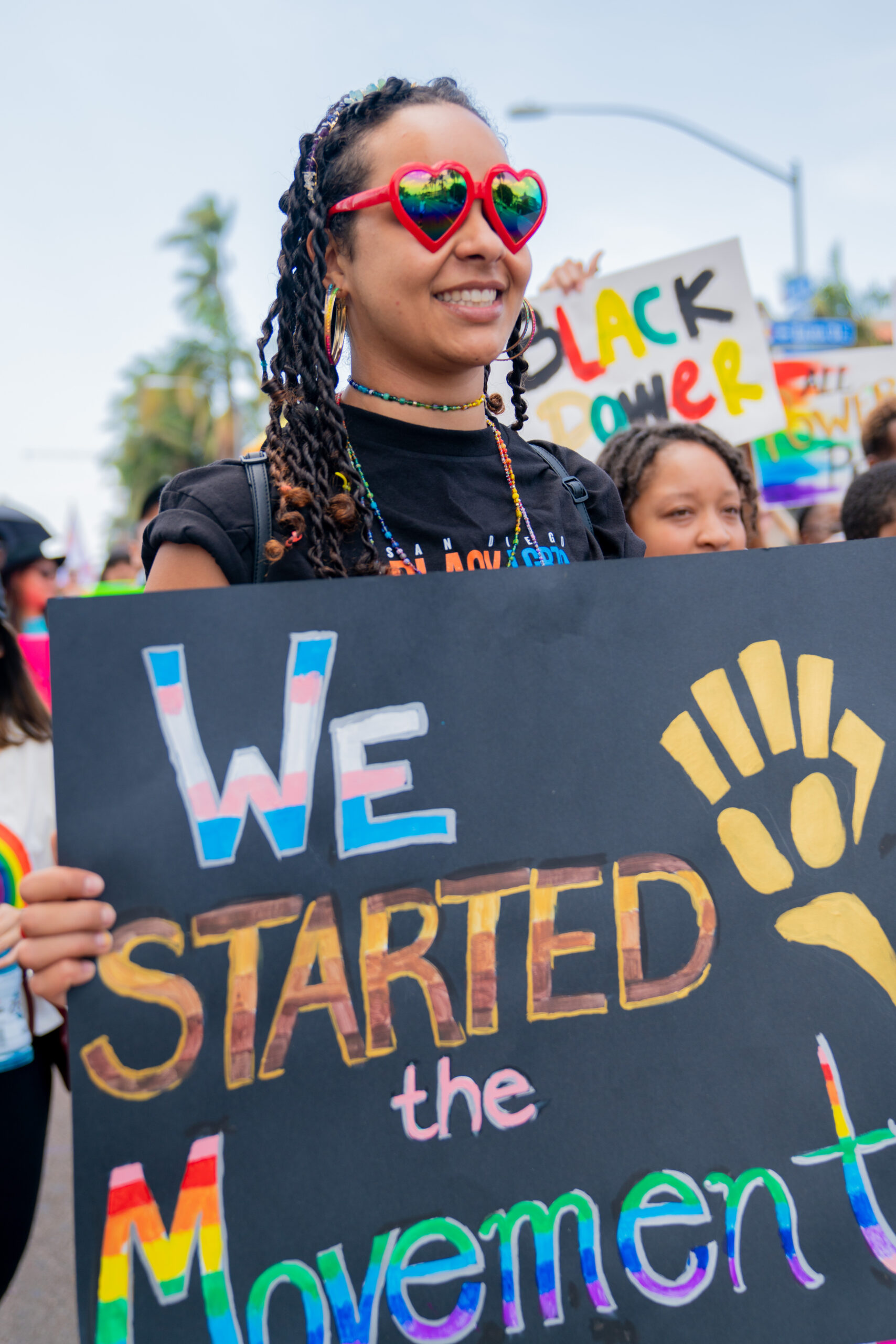 Woman of color in red heart shaped sunglasses and locs holds a sign that says 