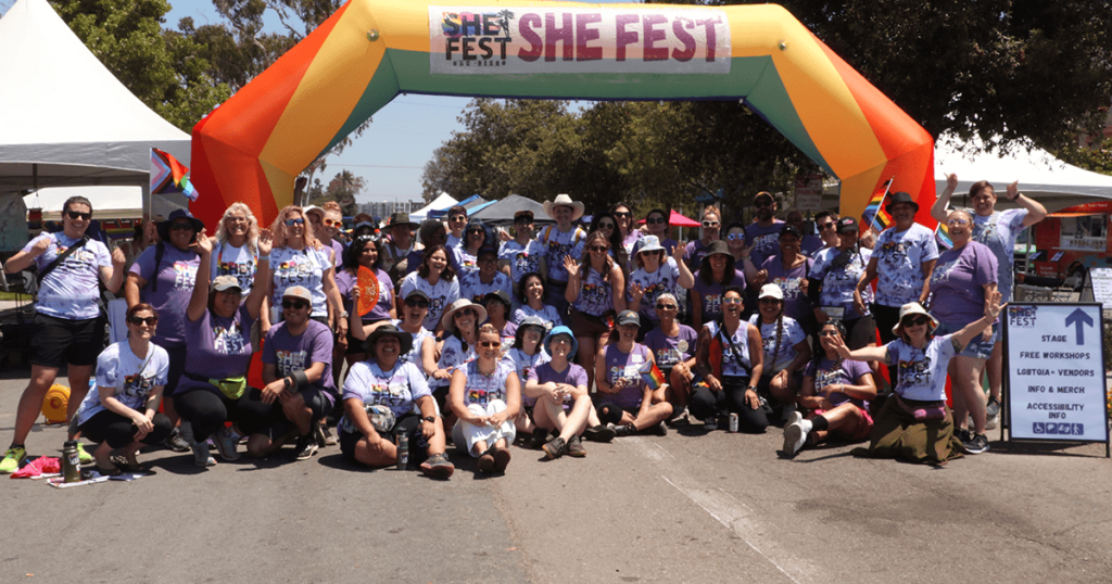 She Fest Planning Committee and volunteers huddled and smiling underneath the She Fest blow up arch.
