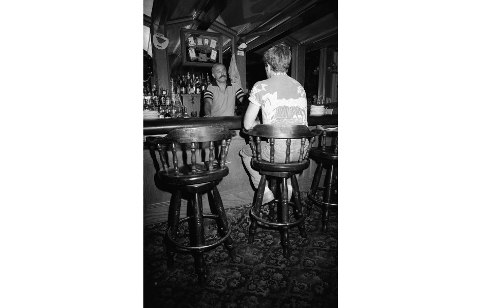 A male bartender with a mustache chats with a patron at the bar